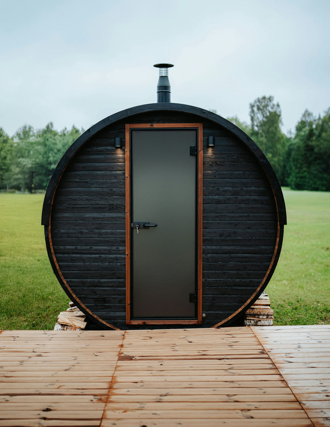 Dark wooden barrel sauna with a glass door and chimney on a wooden deck in a green field