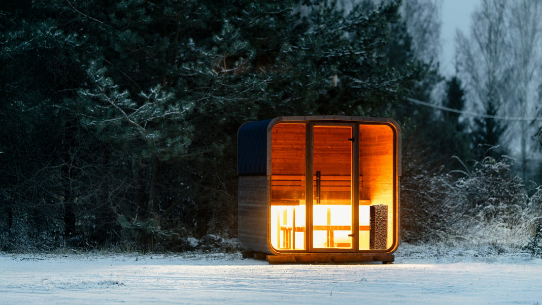 Wooden sauna cabin by a lake at dusk with warm light in the window and smoke from the chimney